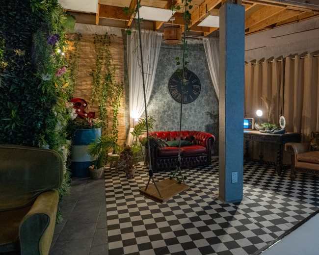 The image shows a stylish indoor space featuring a mix of vintage and modern furniture, including a red leather sofa and a wooden desk, surrounded by plants and decorative elements.