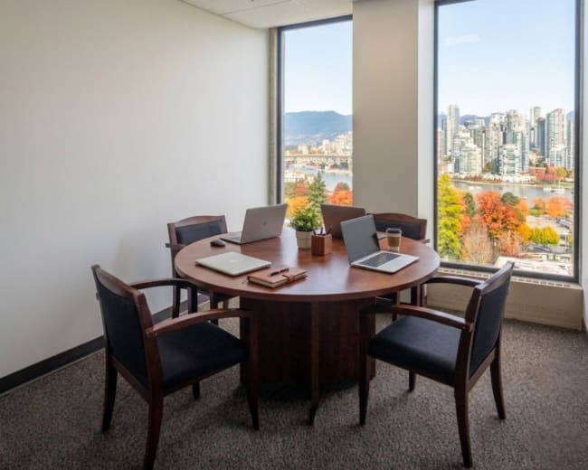 A modern office with a round wooden table and four chairs overlooks a cityscape and mountains through large windows.