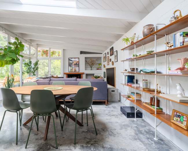 The image shows a bright, modern living space featuring a round wooden dining table surrounded by four green chairs, with a lounge area in the background and a bookshelf against the wall.
