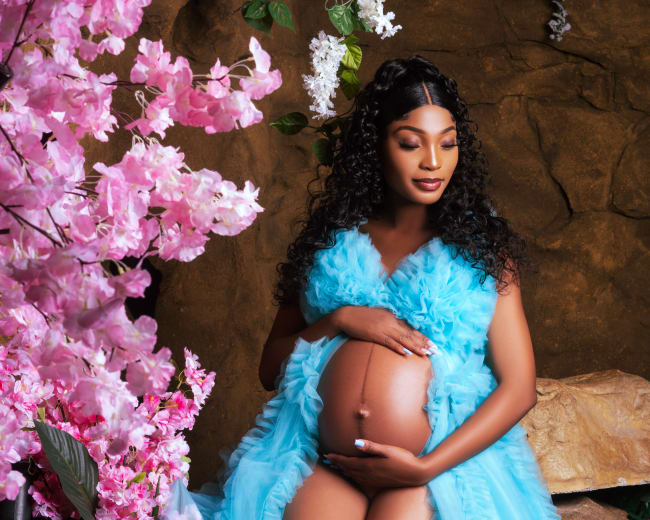 A woman in a blue dress gently holds her pregnant belly while seated among pink flowers and decorative rocks.