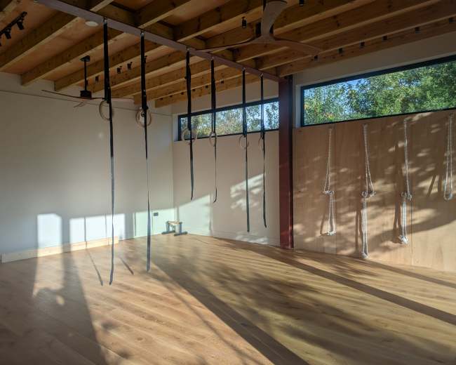 The image depicts a spacious, empty room with wooden floors, exposed beams, and apparatus such as ropes and rings suspended from the ceiling.