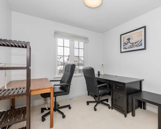 The image shows a bright home office with two black chairs, a wooden desk, a black desk with a drawer, and a shelf against the wall.
