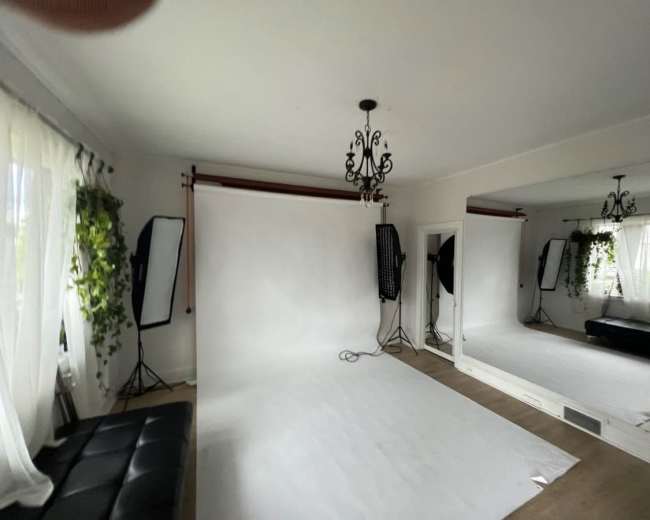 A well-lit studio space features a white backdrop, two softbox lights, a large mirror, and a black sofa.