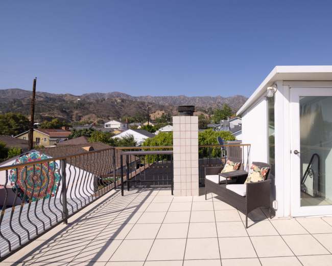 The image shows a rooftop terrace with a seating area, a chimney, and a mountainous backdrop under a clear blue sky.