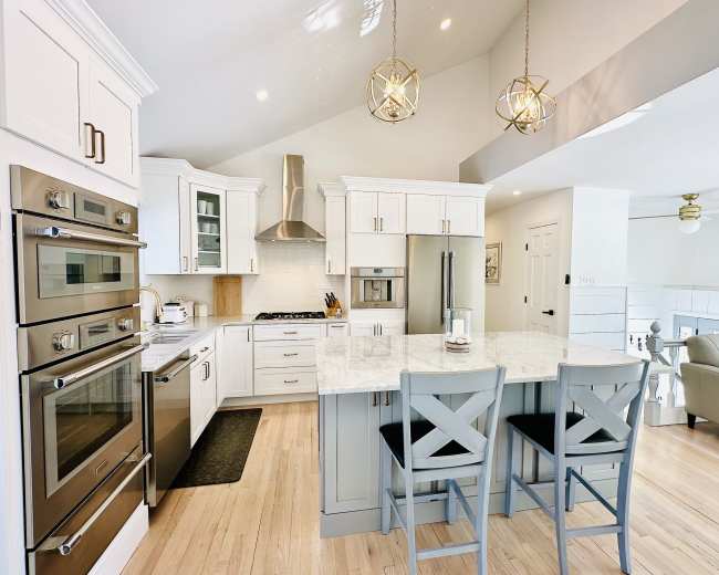 The image shows a modern kitchen with white cabinetry, stainless steel appliances, a marble countertop, and two gray bar stools at the island.