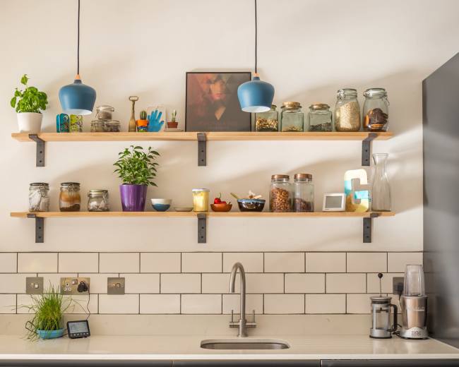 A modern kitchen features gray cabinetry, open wooden shelves filled with jars and plants, and a stainless steel sink.