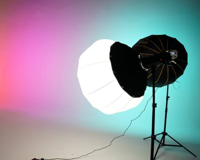 The image shows a photography studio setup with three light sources, including two softboxes and a small spotlight, against a gradient backdrop of pink and blue.
