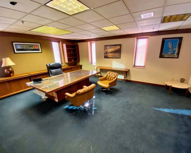 The image shows an office with a large wooden desk, a black leather office chair, two brown armchairs, and framed pictures on the walls.