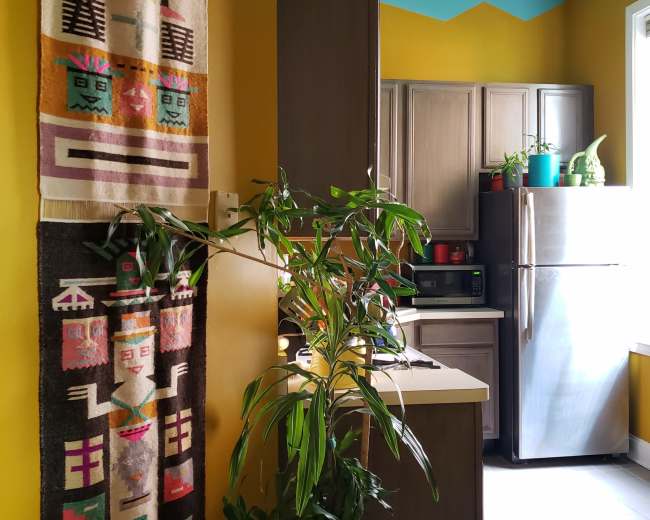 The image shows a brightly colored kitchen featuring yellow walls, a turquoise ceiling, and a refrigerator, with two woven wall hangings and a potted plant visible.