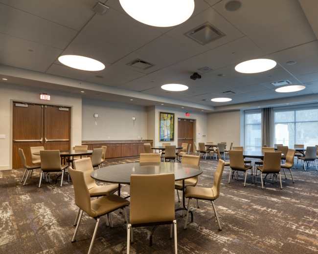 The image shows a spacious, modern conference room with several round tables and chairs arranged for meetings or events, illuminated by circular pendant lights.