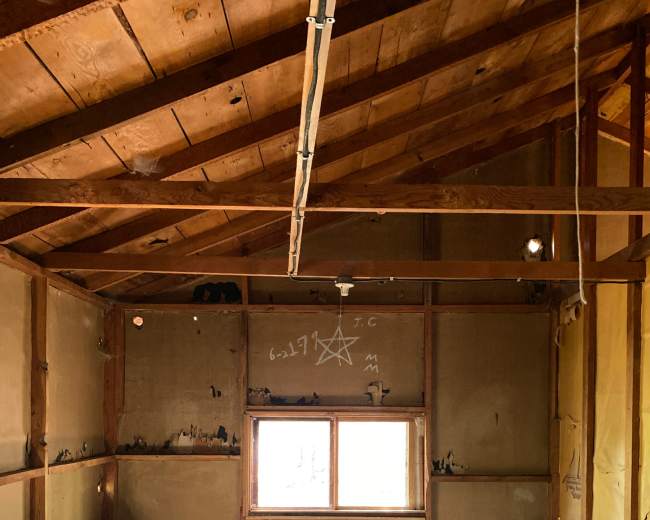 An empty room with wooden benches and a table beneath a slanted roof, featuring a window and markings on the walls.