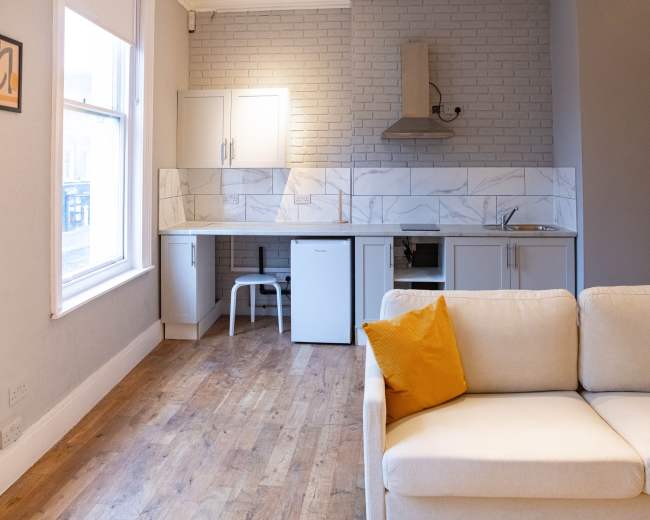The image shows a modern, minimalist living space featuring a small kitchen area with white cabinetry, a stove hood, and a refrigerator, alongside a light-colored sofa with yellow throw pillows.
