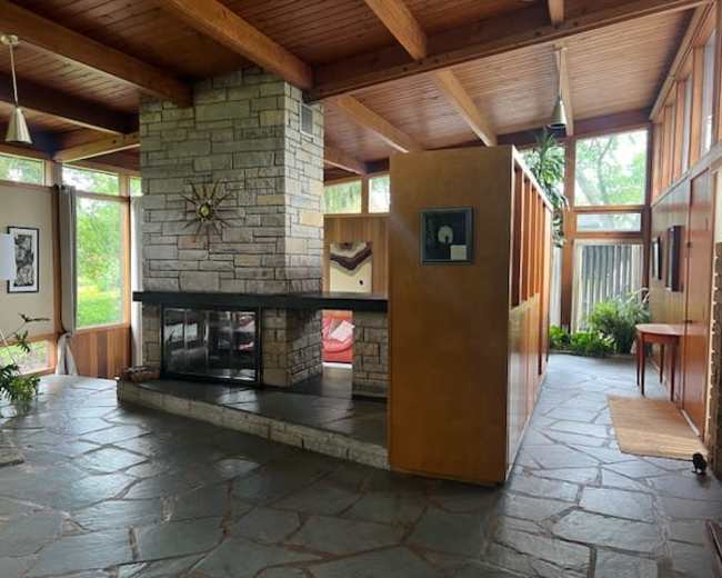 The image depicts a modern interior space featuring a stone fireplace, wooden beams, and large windows allowing natural light to enter.