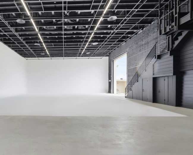 The image shows an empty, open studio space with a white backdrop, concrete flooring, and a staircase leading to a second level.