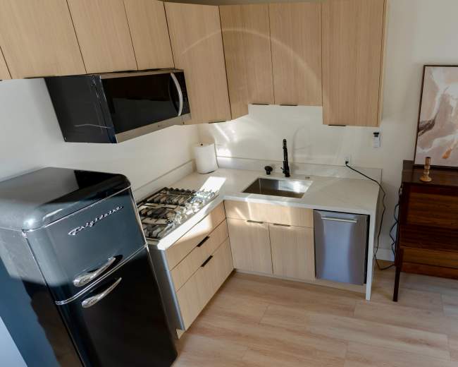 The image shows a modern kitchen featuring light wood cabinetry, a black retro refrigerator, a silver sink, and decorative artwork on the wall.