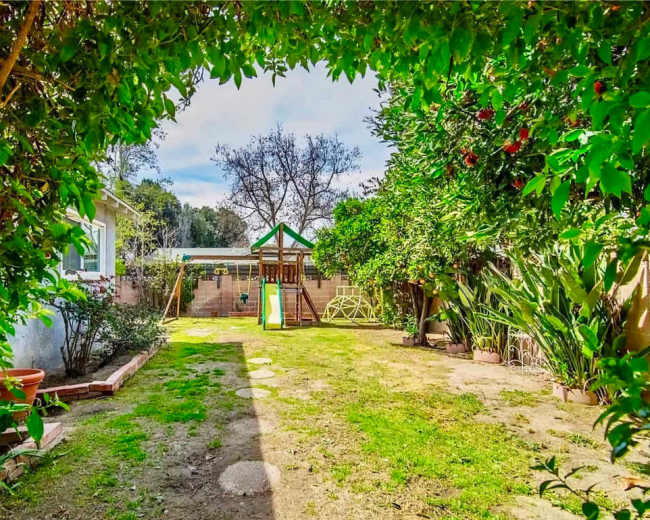 The image shows a backyard with a grassy area, a children’s playset, and various trees and shrubs.