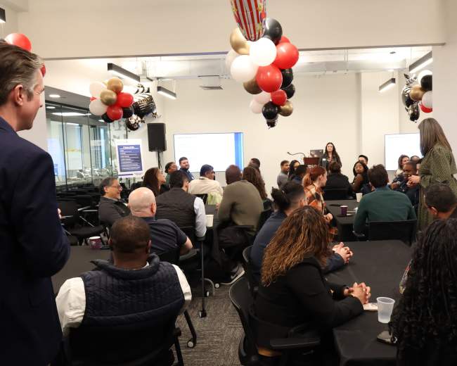 A group of people sits at round tables in a conference room, engaged in discussion, while balloons and decorations hang from the ceiling.