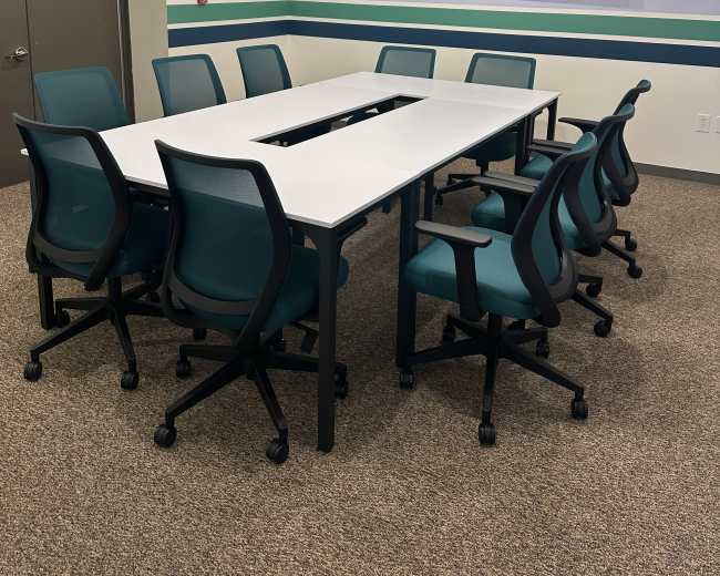 The image shows a modern conference room featuring a long rectangular table surrounded by eight teal chairs, with a wall-mounted television and striped wall decor.