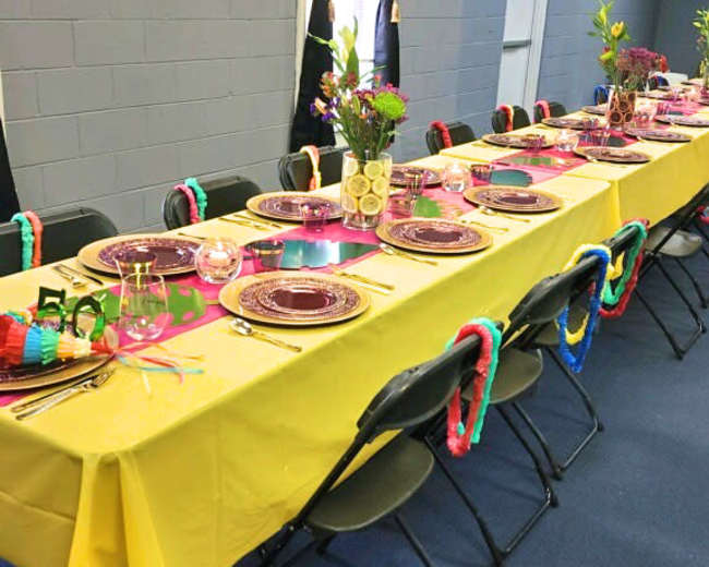 A long table is set with yellow tablecloths and adorned with plates, glasses, and floral arrangements, ready for an event.