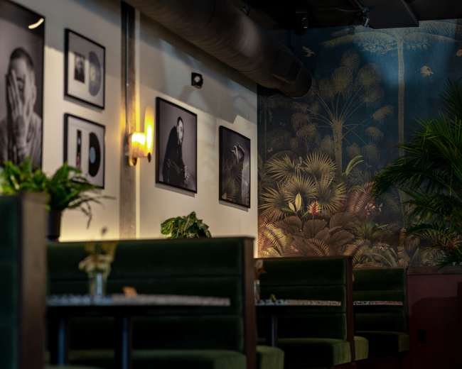The image shows a stylish restaurant interior featuring green booths, framed black-and-white photographs on the walls, and a large mural of tropical plants.