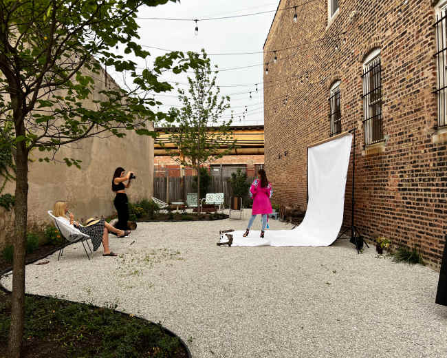 A woman in a pink outfit poses in front of a white backdrop while another woman takes her photograph in a gravel yard surrounded by brick walls.