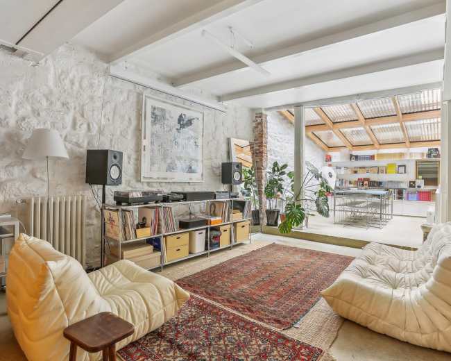 The image shows a contemporary living space with white stone walls, a seating area featuring large cushions, and an open kitchen visible in the background.
