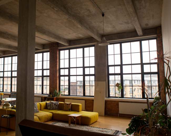 The image shows an industrial-style living room featuring large windows, a yellow sofa, and a wooden dining table.