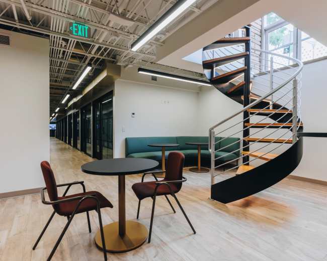 The image shows a modern office space featuring a spiral staircase, circular table with two chairs, and glass-walled meeting rooms.
