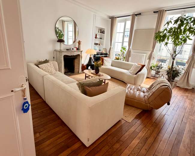 The image shows a bright living room featuring two white sofas, a round mirror, a small fireplace, and indoor plants arranged around a wooden floor.