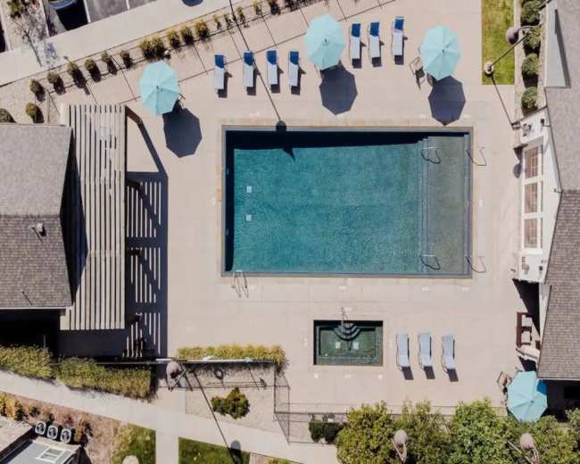 An aerial view of a rectangular swimming pool surrounded by lounge chairs and umbrellas, with a small fountain nearby.