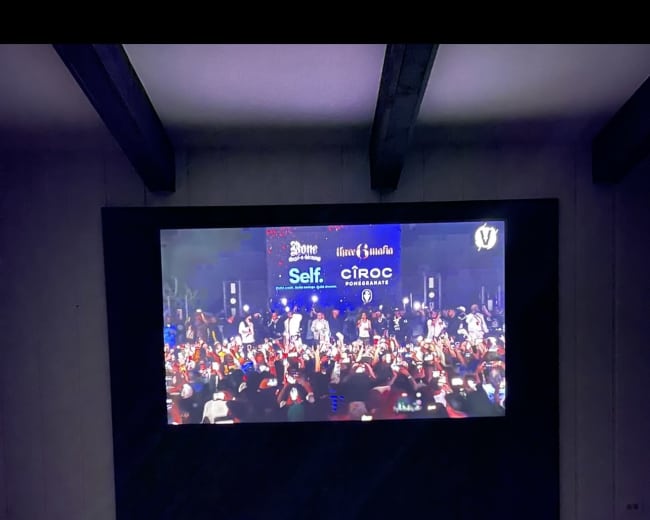 A large projector screen displays a lively event with a crowd gathered for a performance in a modern room with a red carpet.