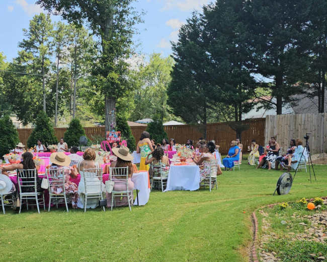 A garden party is taking place with guests seated at tables set up on a grassy area, surrounded by trees and a wooden fence.