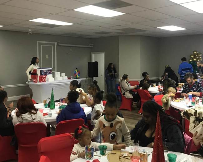 A group of people gathered around tables decorated for a festive event in a room with balloons and a speaker.