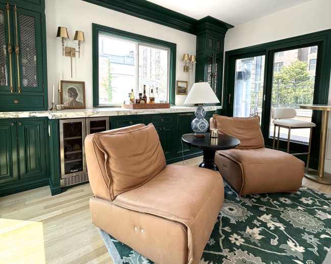 The image shows a stylish living room with two tan lounge chairs, a small round table, and green cabinetry, illuminated by natural light from large windows.