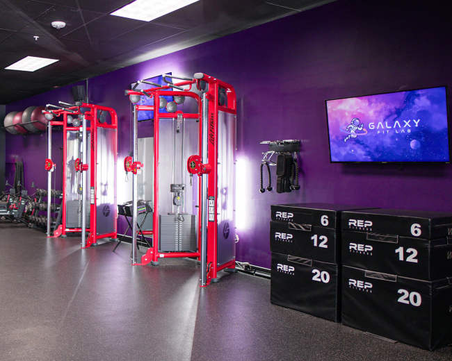 The image shows a fitness studio with workout equipment including cable machines, weights, and storage boxes against a purple wall.
