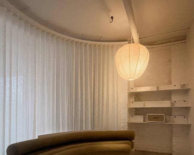 The image features a modern interior with a curved brown sofa, a white round coffee table, and a large paper lantern light, set against a backdrop of sheer curtains and white walls.
