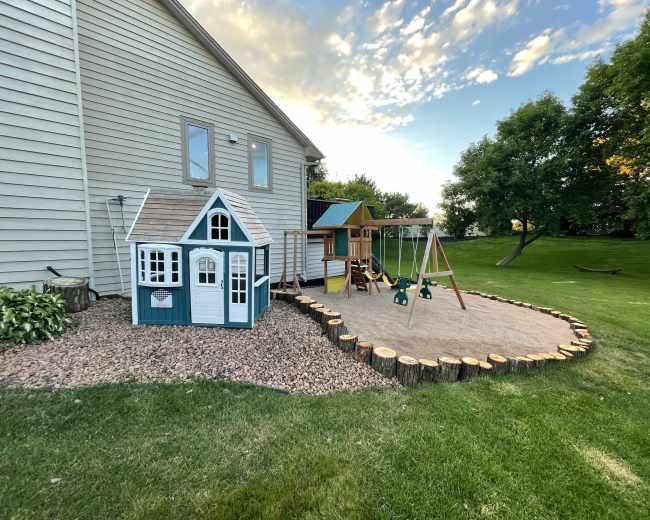 A backyard play area featuring a small blue and white playhouse, a wooden swing set, and a sandy playing surface surrounded by tree stumps.