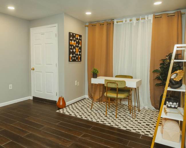 The image shows a small room with a table and chairs, a plant, and a ladder shelf against the wall, featuring light brown and white curtains.