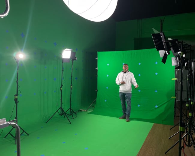 A man stands in front of a green screen setup, surrounded by studio lights and equipment.