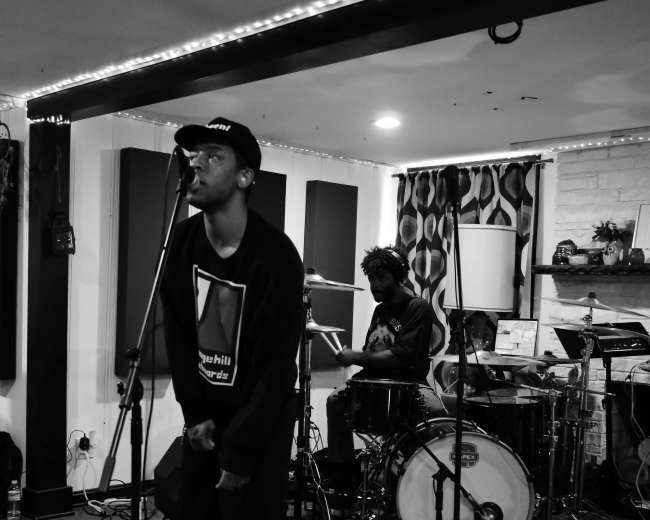 A musician stands at a microphone while a drummer plays in a dimly lit studio.