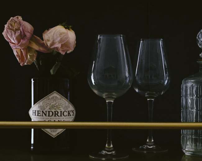 A black background with a bottle of Hendrick's gin, two wine glasses, a glass decanter, and a small vase of dried roses.