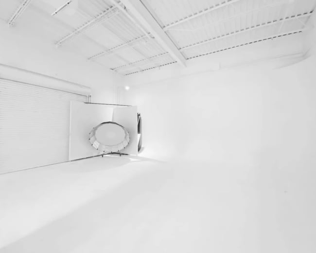 A minimalistic, white interior space with a large circular mirror and a closed roll-up door against a wall.