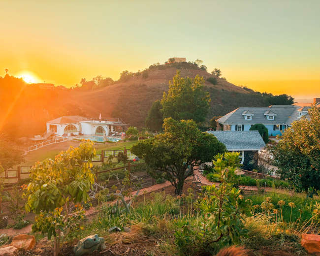 The image shows a scenic view of a hillside at sunset, with houses and gardens in the foreground and a bright orange sky in the background.