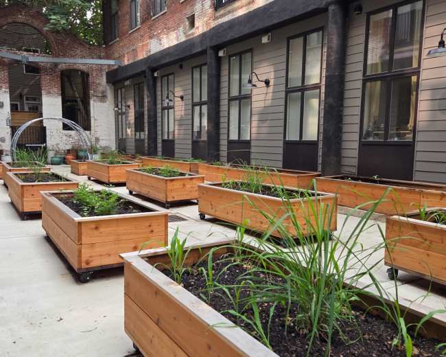 The image shows a courtyard with wooden planter boxes filled with soil and plants, bordered by brick and wooden buildings.