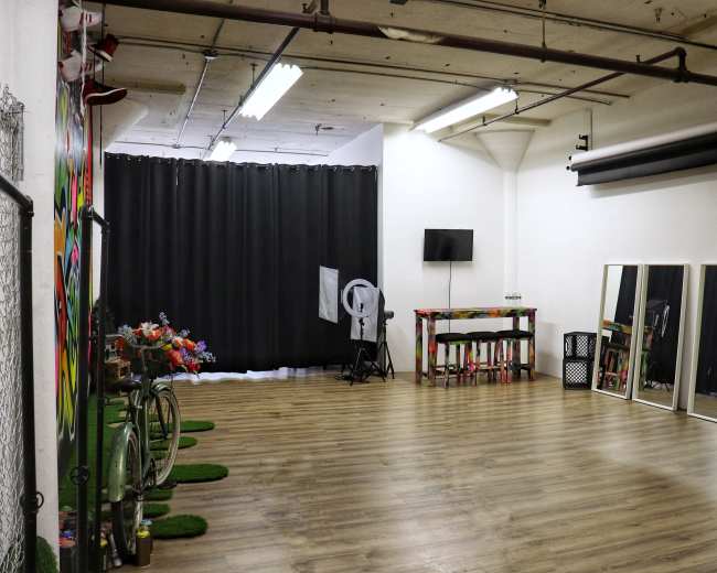A spacious room with wooden flooring, featuring a black curtain at one end, a table with colorful decorations, mirrors, and a television mounted on the wall.