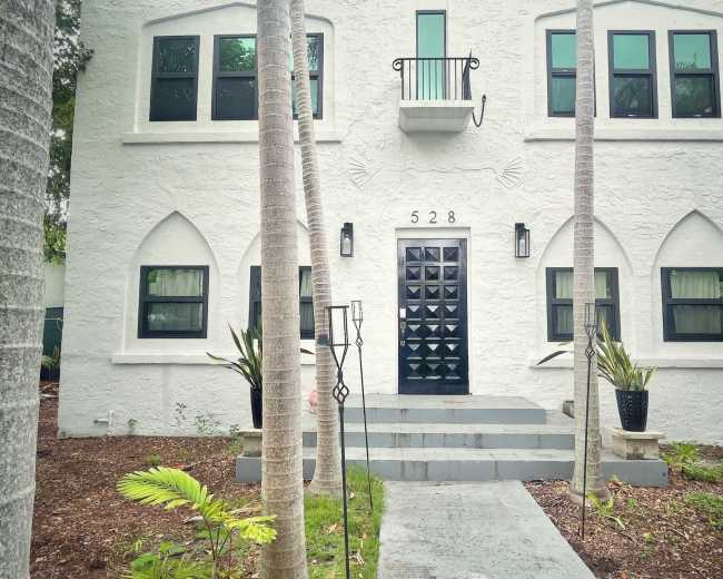 The image features a two-story white building with an ornate black door, flanked by palm trees and a pathway leading to the entrance.