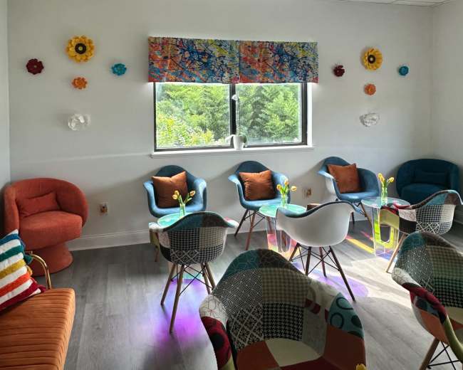 The image shows a brightly lit room decorated with colorful furniture, including chairs and a sofa, along with flower-shaped wall decor and large windows that let in natural light.