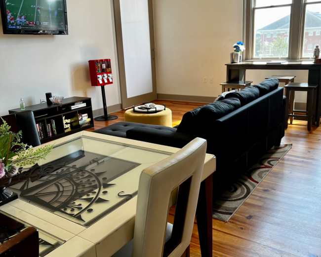 A modern living space features a dining table with a decorative design, a black sofa, a TV displaying a sports game, and a large window illuminating the room.