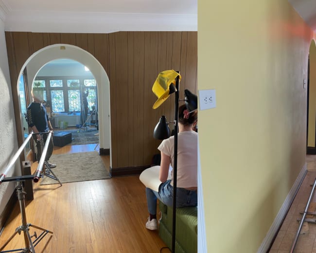Two sections of a film set: one side features a person sitting near a camera setup in a room, while the other side shows a hallway with a track for filming.
