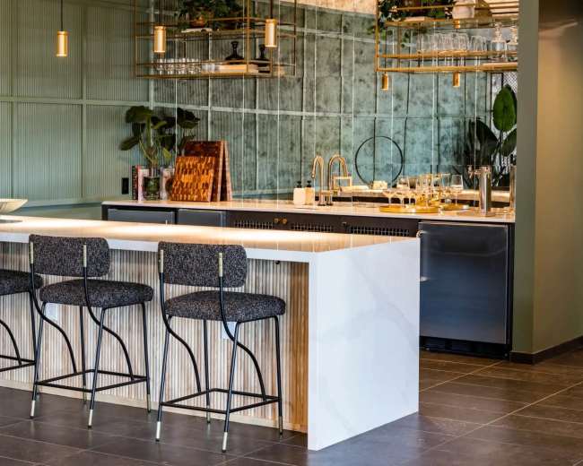 The image shows a modern kitchen with a bar area featuring four chairs, a countertop, and sleek cabinetry.